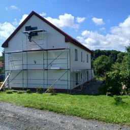 Expres-Bud Krzysztof Kleban - Dwupiętrowy dom z białymi ścianami i czerwonym dachem w trakcie malowania elewacji. Na ścianie rusztowanie, na którym stoi pracownik. Błękitne niebo z chmurami w tle.