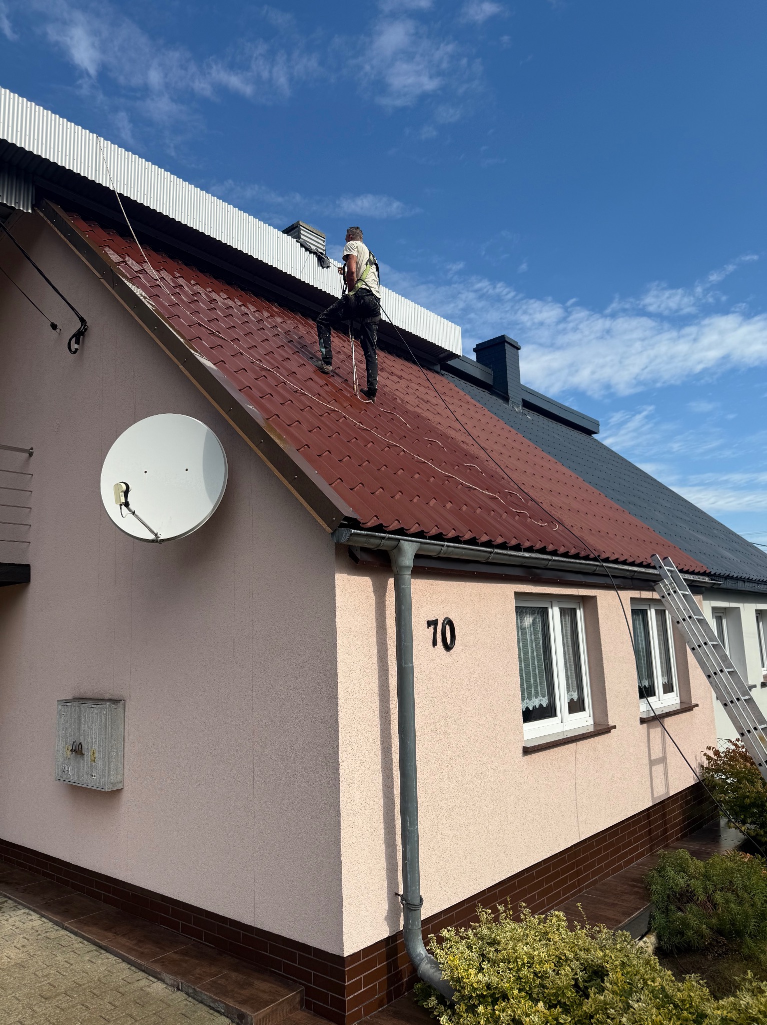 Pracownik w uprzęży myje ciśnieniowo czerwoną, błyszczącą po umyciu część dachu domu z jasną elewacją. Widoczny fragment dachu w kolorze grafitowym i drabina oparta o ścianę.