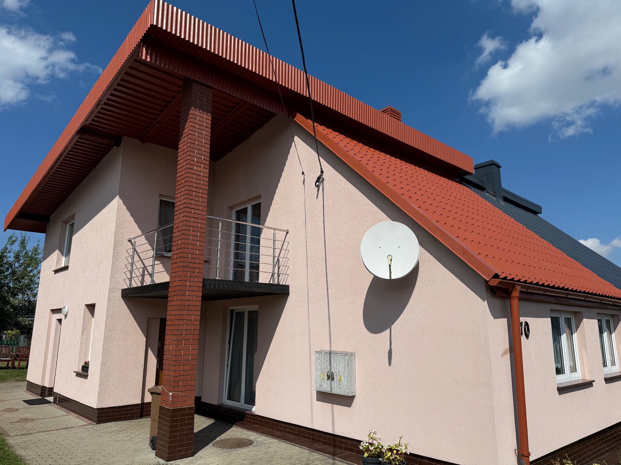 Jasnoróżowy dom z czerwonym dachem i ceglanymi kolumnami balkonu, widziany w słoneczny dzień. Widoczna antena satelitarna i numer domu.