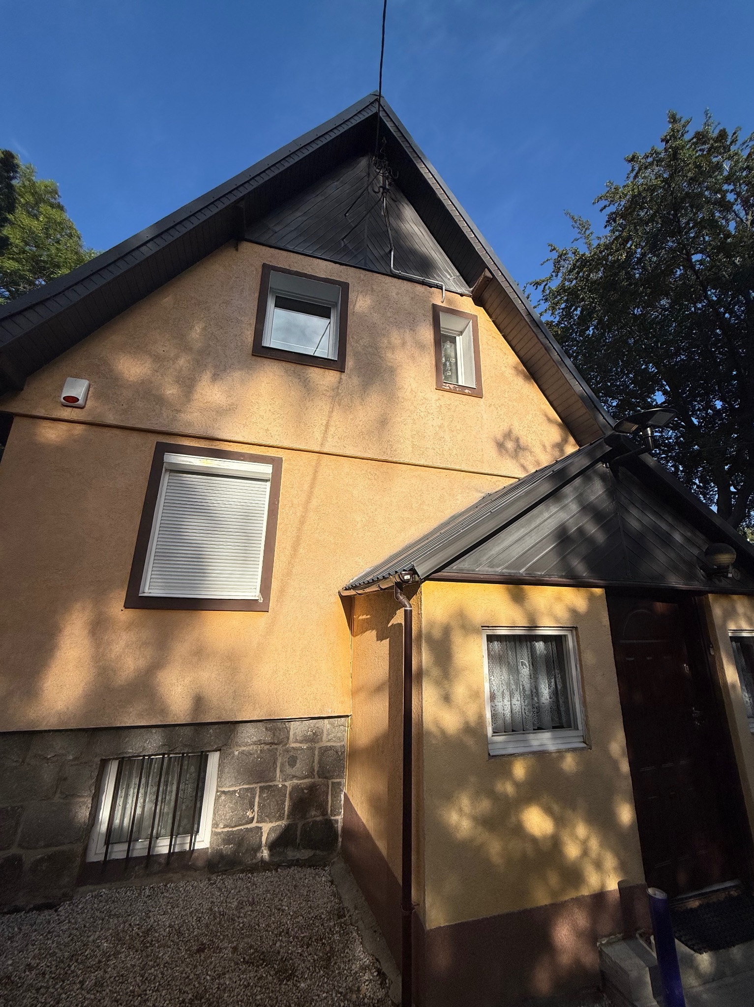 Ukończona elewacja domu z brązowym dachem i oknami z roletami, widziana pod kątem. Część elewacji wykończona kamieniem. Na ścianie czujnik alarmowy.
