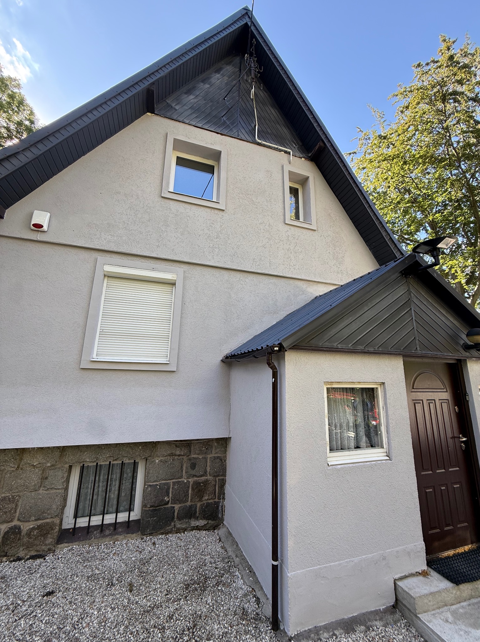 Odnowiona elewacja domu z szarą fasadą, ciemnym dachem i brązowymi drzwiami wejściowymi. Widoczne okna z roletami i kratami. Ujęcie z dołu, podkreślające geometrię budynku.