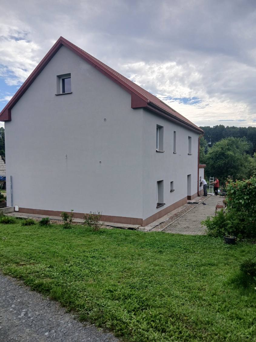Jasnoszary dom z czerwoną dachówką po malowaniu elewacji. Dwóch pracowników przy cokole. Widoczne drzewa w tle i fragment trawnika. Ujęcie z dołu.