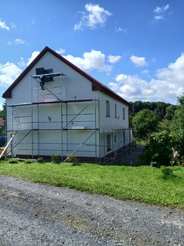 Dwupiętrowy dom z białymi ścianami i czerwonym dachem w trakcie malowania elewacji. Na ścianie rusztowanie, na którym stoi pracownik. Błękitne niebo z chmurami w tle.