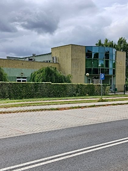 Dwukondygnacyjny budynek z zaokrągloną, przeszkloną fasadą, otoczony zielenią i chodnikiem. Widoczne elementy architektoniczne i otoczenie miejskie.