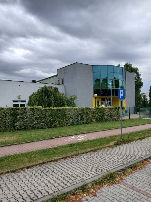 Szary budynek z żółtymi akcentami i niebieskimi szybami, widoczny z chodnika z kostki brukowej. Na tle pochmurne niebo. Widoczny znak parkingu.