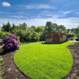 4 Pory Roku - Zadbany trawnik z rabatą kwiatową i drewnianą altaną w tle, otoczony zielenią drzew, pod błękitnym niebem z chmurami. Widok z poziomu gruntu.