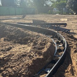 4 Pory Roku - Montaż obrzeży trawnikowych z tworzywa na przygotowanym podłożu z piasku. Obrzeża ułożone w łuk, w tle zieleń drzew i krzewów.