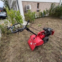 4 Pory Roku - Czerwony glebogryz na trawniku przed nowoczesnym domem, w tle zaparkowane samochody. Widoczny teren po przekopaniu.