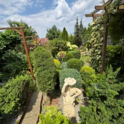 Ogród z rzeźbą kobiety w kapeluszu, różnorodne krzewy i drzewa iglaste, drewniana pergola z pnączami, ścieżka z kamieniami. Słoneczny dzień, błękitne niebo z obłokami.