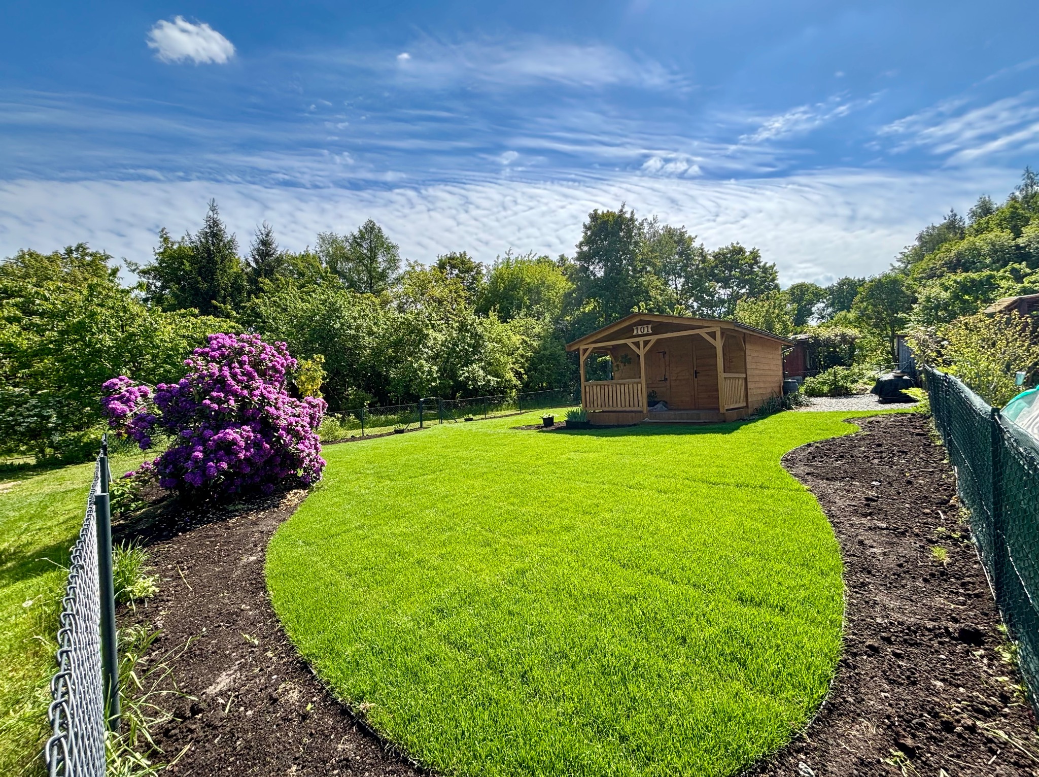 Zadbany trawnik z rabatą kwiatową i drewnianą altaną w tle, otoczony zielenią drzew, pod błękitnym niebem z chmurami. Widok z poziomu gruntu.