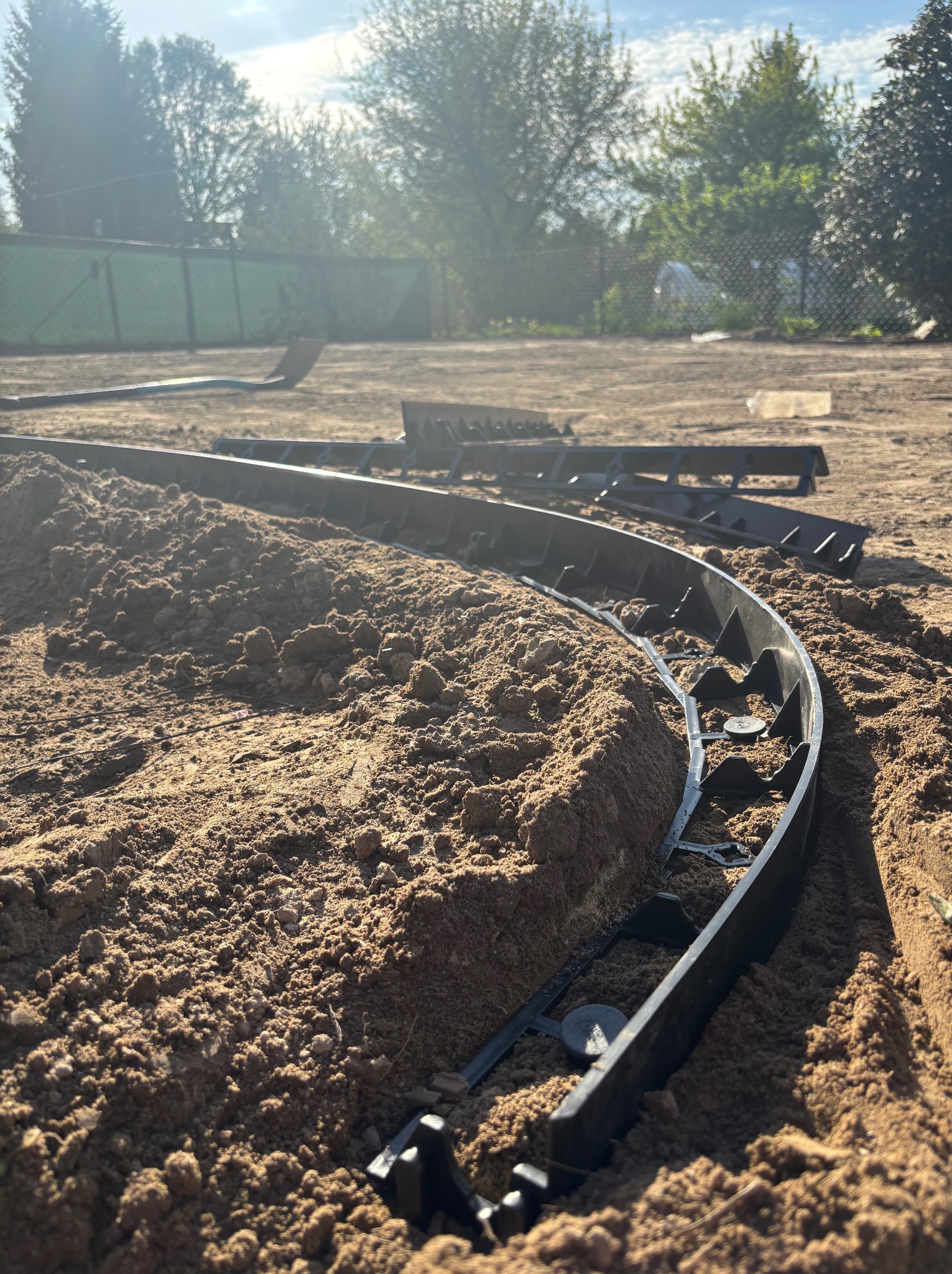 Montaż obrzeży trawnikowych z tworzywa na przygotowanym podłożu z piasku. Obrzeża ułożone w łuk, w tle zieleń drzew i krzewów.