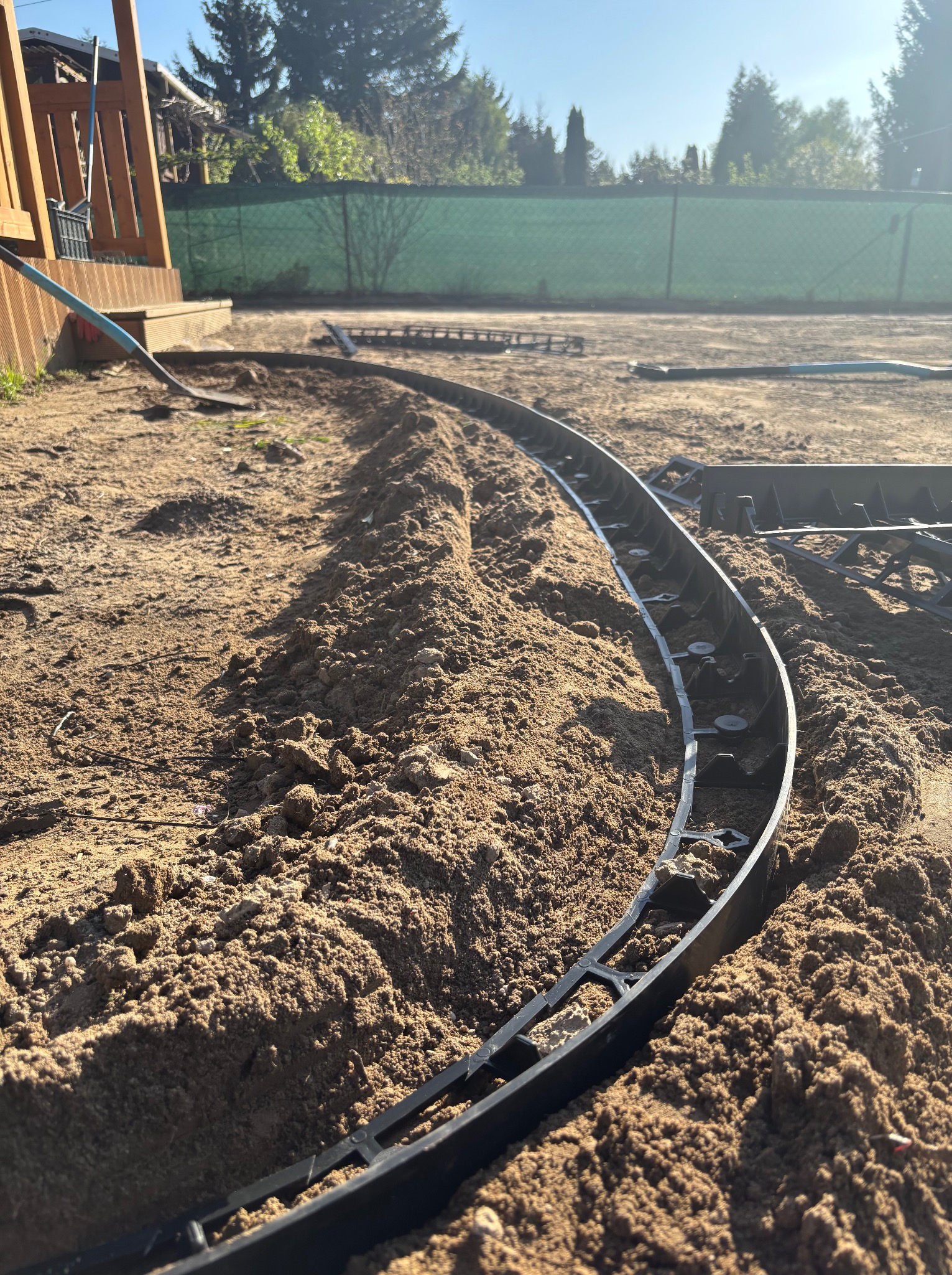 Krawężnik trawnikowy z tworzywa sztucznego ułożony w piasku, wyznaczający ścieżkę w ogrodzie. W tle zielone ogrodzenie i elementy drewnianej konstrukcji.