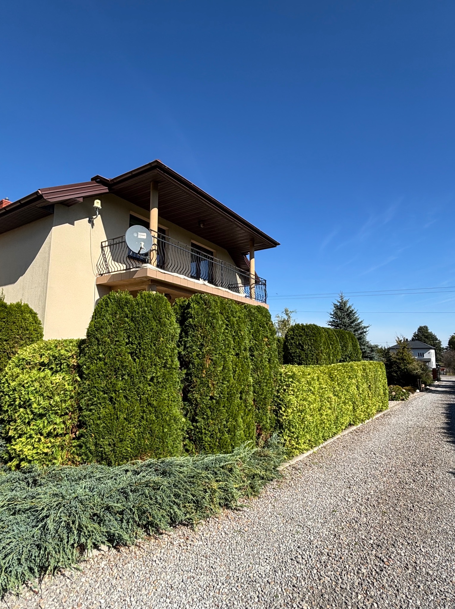 Dom jednorodzinny z beżową elewacją i balkonem, otoczony równo przyciętym żywopłotem z tui, widok z żwirowej ścieżki w słoneczny dzień.