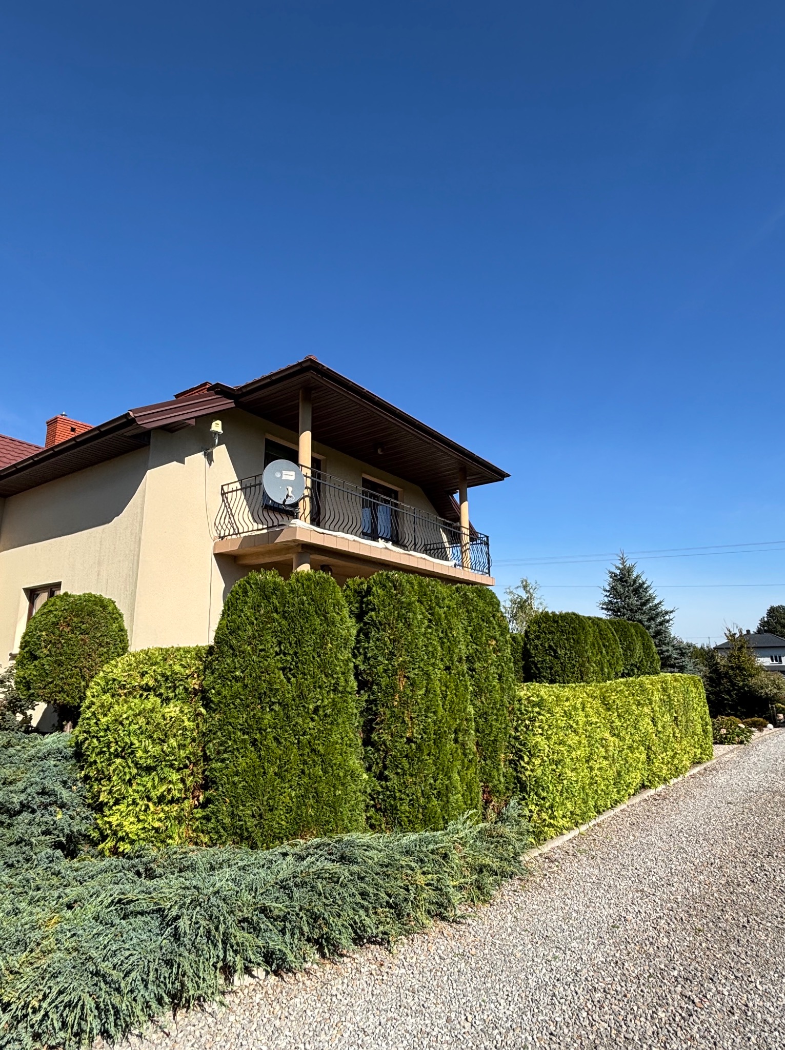 Ogród z formowanymi krzewami i żywopłotem, widok na dom z balkonem i żwirową ścieżką, słoneczny dzień.