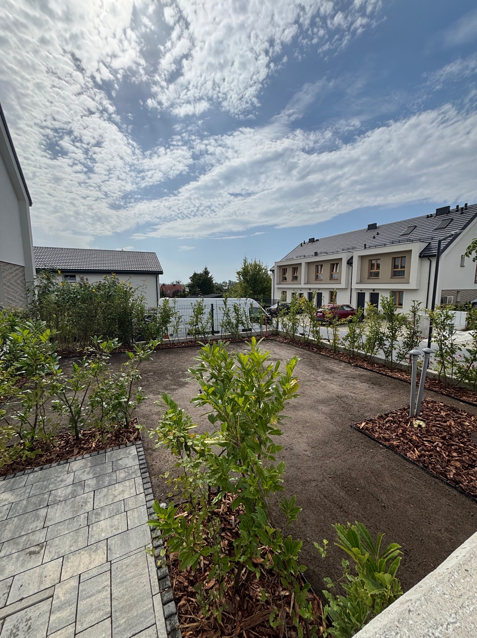 Przygotowany teren pod trawnik z nasadzeniami krzewów wzdłuż ogrodzenia i wykończonym obrzeżem z kory. Widok na nowoczesne budynki w tle.