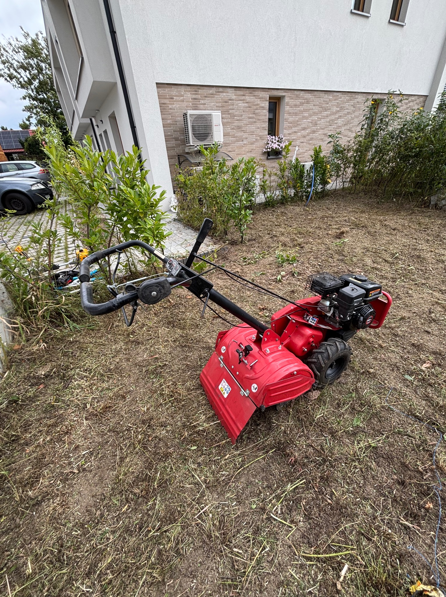Czerwony glebogryz na trawniku przed nowoczesnym domem, w tle zaparkowane samochody. Widoczny teren po przekopaniu.