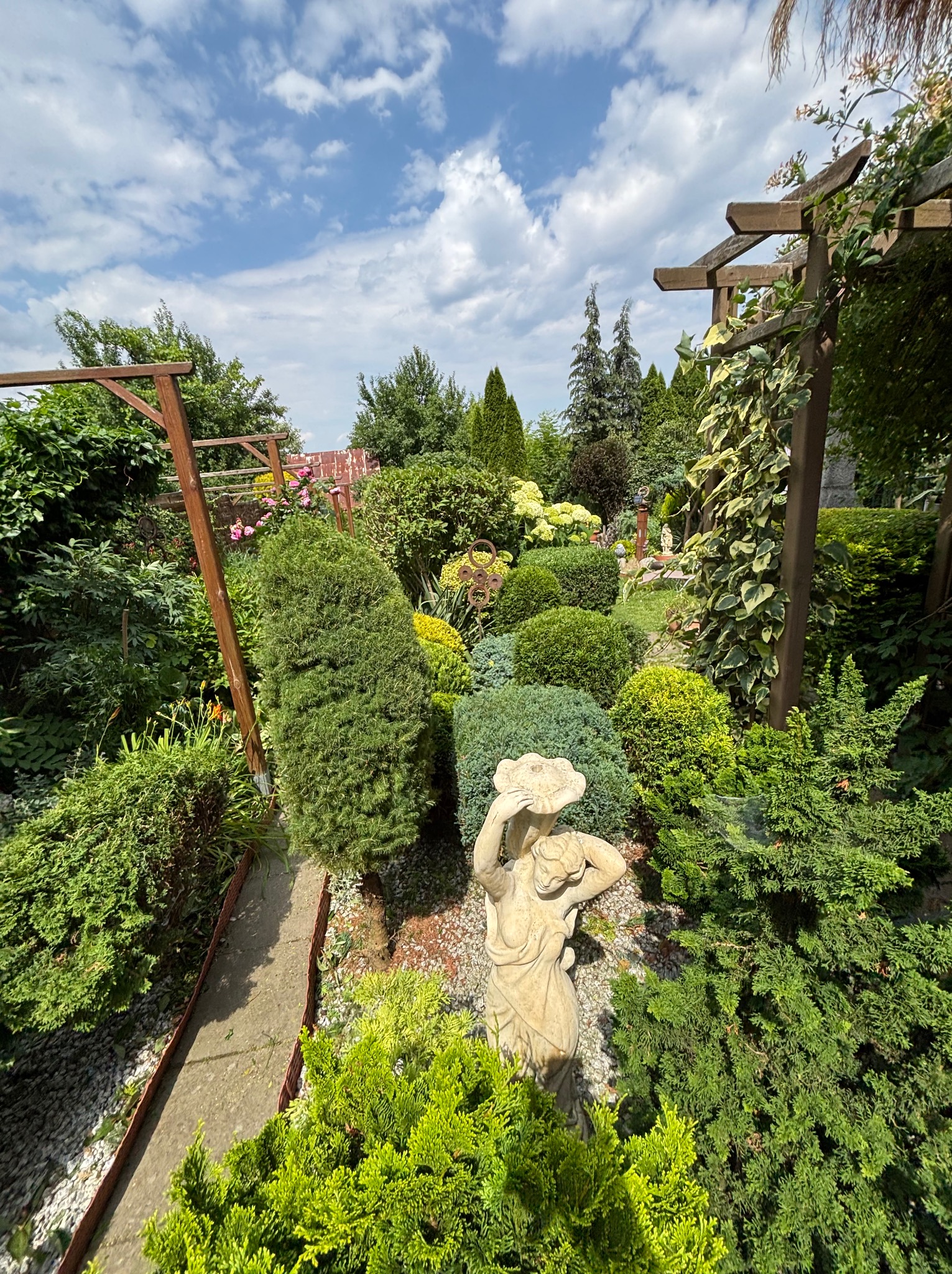 Ogród z rzeźbą kobiety w kapeluszu, różnorodne krzewy i drzewa iglaste, drewniana pergola z pnączami, ścieżka z kamieniami. Słoneczny dzień, błękitne niebo z obłokami.