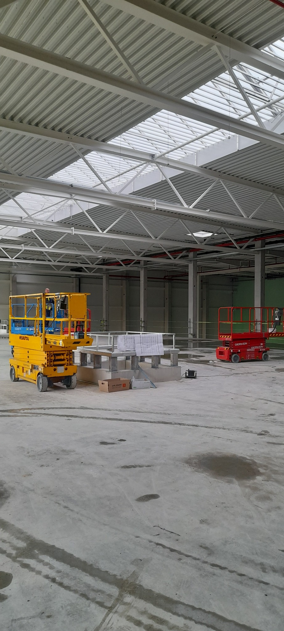 Wnętrze hali w budowie z podnośnikami koszowymi: żółtym i czerwonym. Widoczne plany na podwyższeniu. Biała konstrukcja dachu z oknami. Betonowa podłoga.