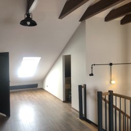 OMITEN SPÓŁKA Z OGRANICZONĄ ODPOWIEDZIALNOŚCIĄ - Poddasze po remoncie: drewniane belki, panele podłogowe, jasne ściany, schody z metalowymi balustradami i drewnianym poręczą. Widok na okno dachowe i oświetlenie.
