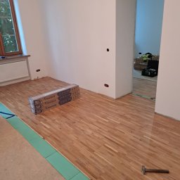 OMITEN SPÓŁKA Z OGRANICZONĄ ODPOWIEDZIALNOŚCIĄ - Układanie parkietu Barlinek w pokoju. Widoczny fragment podkładu, paczki paneli, młotek. Częściowo ułożona podłoga w jasnym odcieniu drewna.