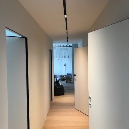 OMITEN SPÓŁKA Z OGRANICZONĄ ODPOWIEDZIALNOŚCIĄ - Biały korytarz po remoncie z drewnianą podłogą, czarnymi obramowaniami drzwi i nowoczesnym oświetleniem sufitowym. Widok na wnętrze z workiem i kartonami.