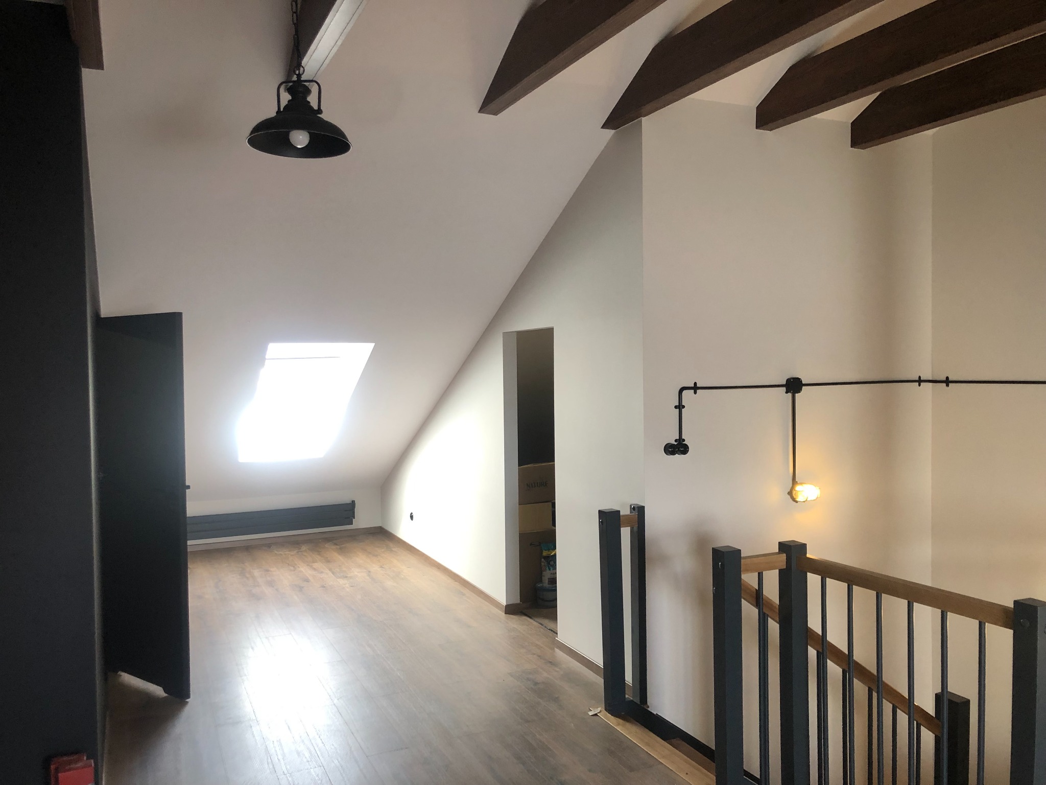 Poddasze po remoncie: drewniane belki, panele podłogowe, jasne ściany, schody z metalowymi balustradami i drewnianym poręczą. Widok na okno dachowe i oświetlenie.