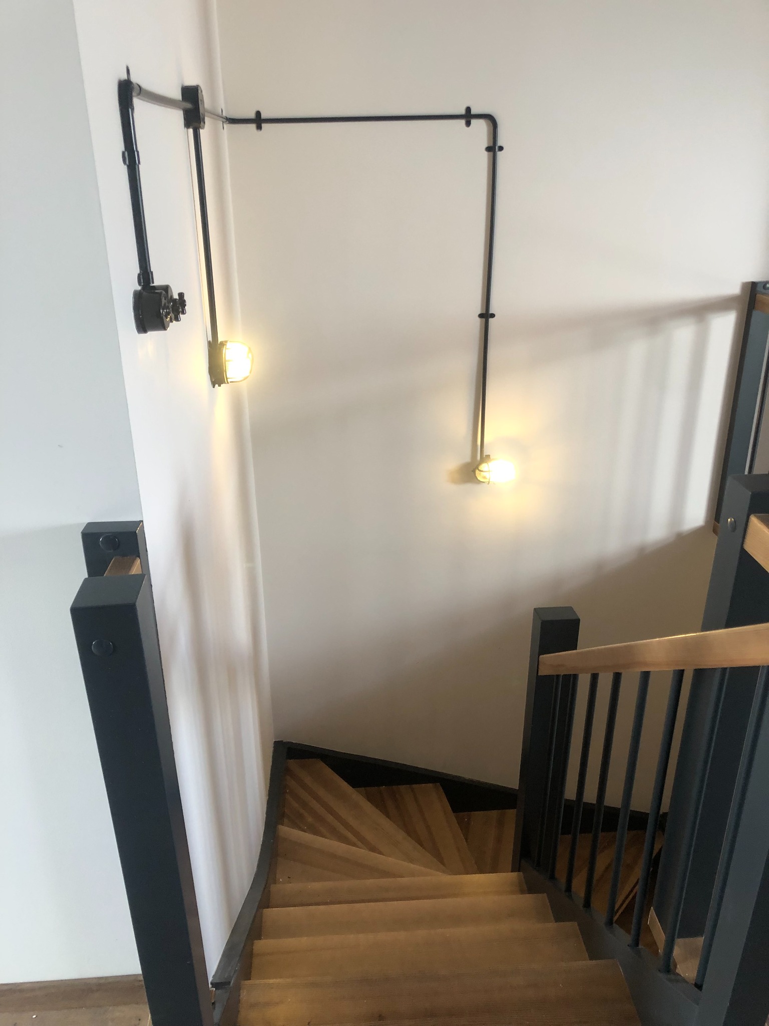 Klatka schodowa z drewnianymi stopniami i czarną balustradą. Na ścianie industrialne oświetlenie z czarnymi rurami i dwoma punktami świetlnymi. Widok z góry.