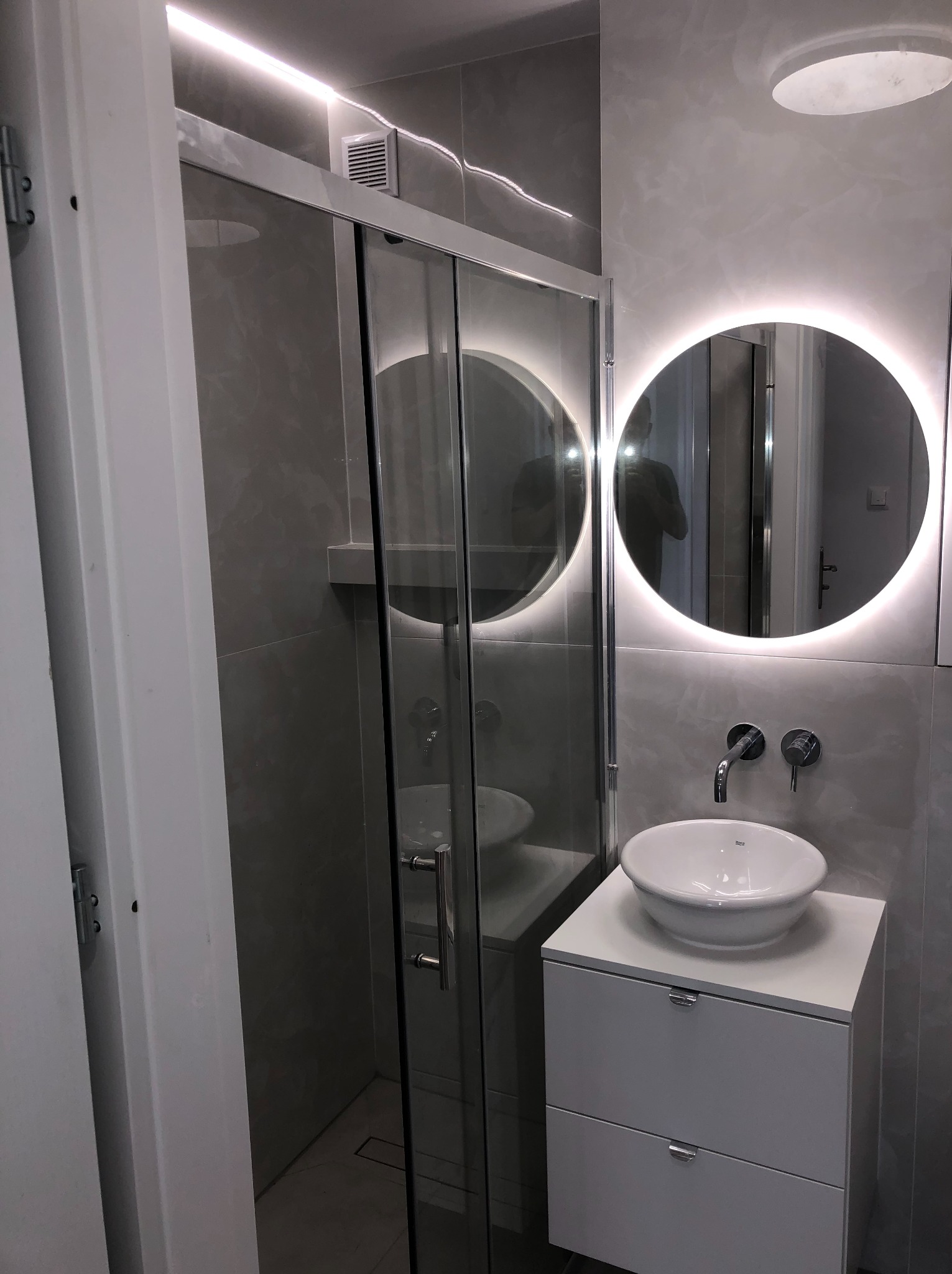 Wykończona łazienka w szarościach: kabina prysznicowa ze szklanymi drzwiami, okrągłe lustro z podświetleniem LED, umywalka nablatowa na białej szafce.