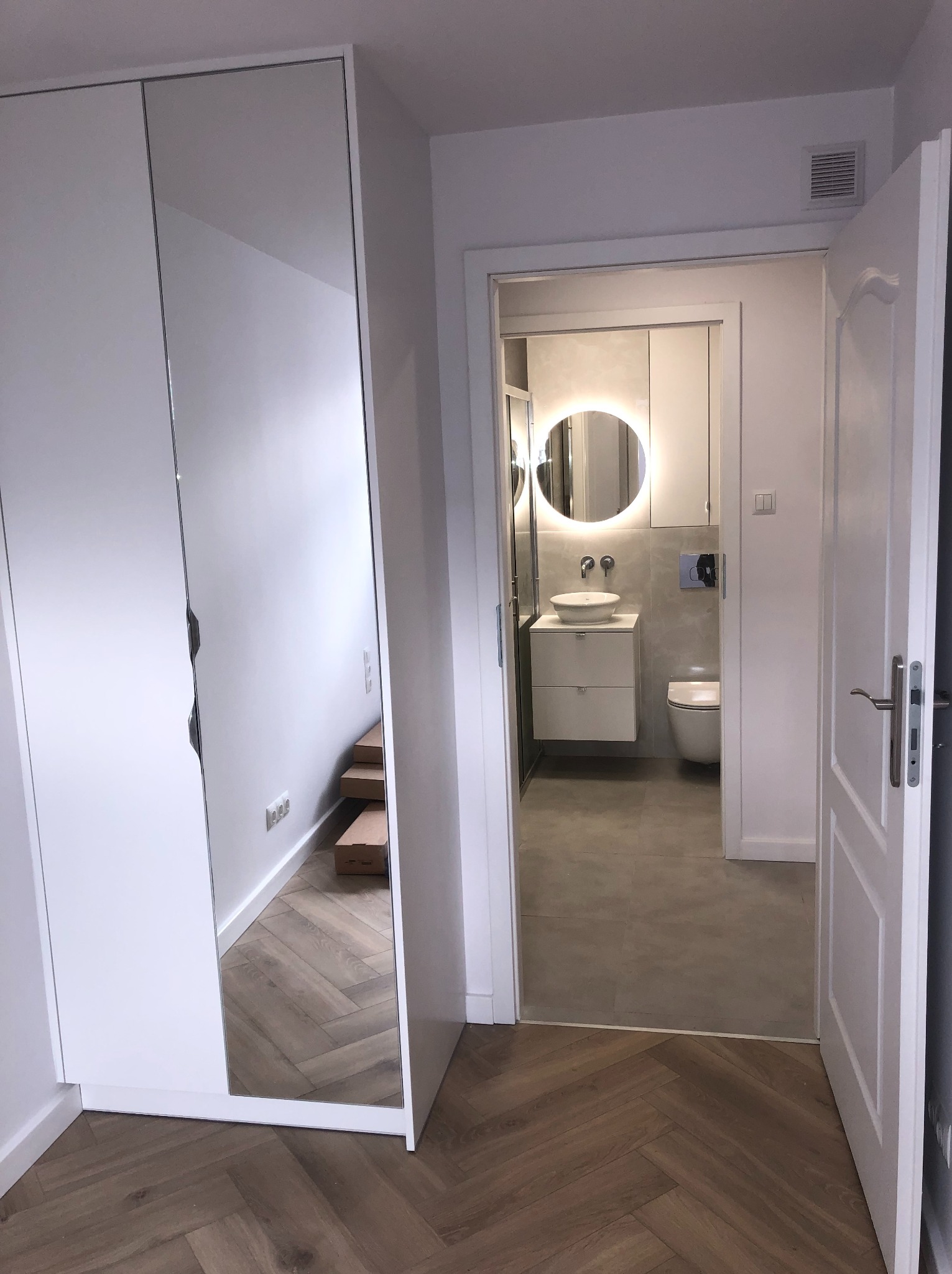 Nowoczesne wnętrze: biała szafa z lustrem, parkiet w jodełkę i widok na wyremontowaną łazienkę z okrągłym lustrem.