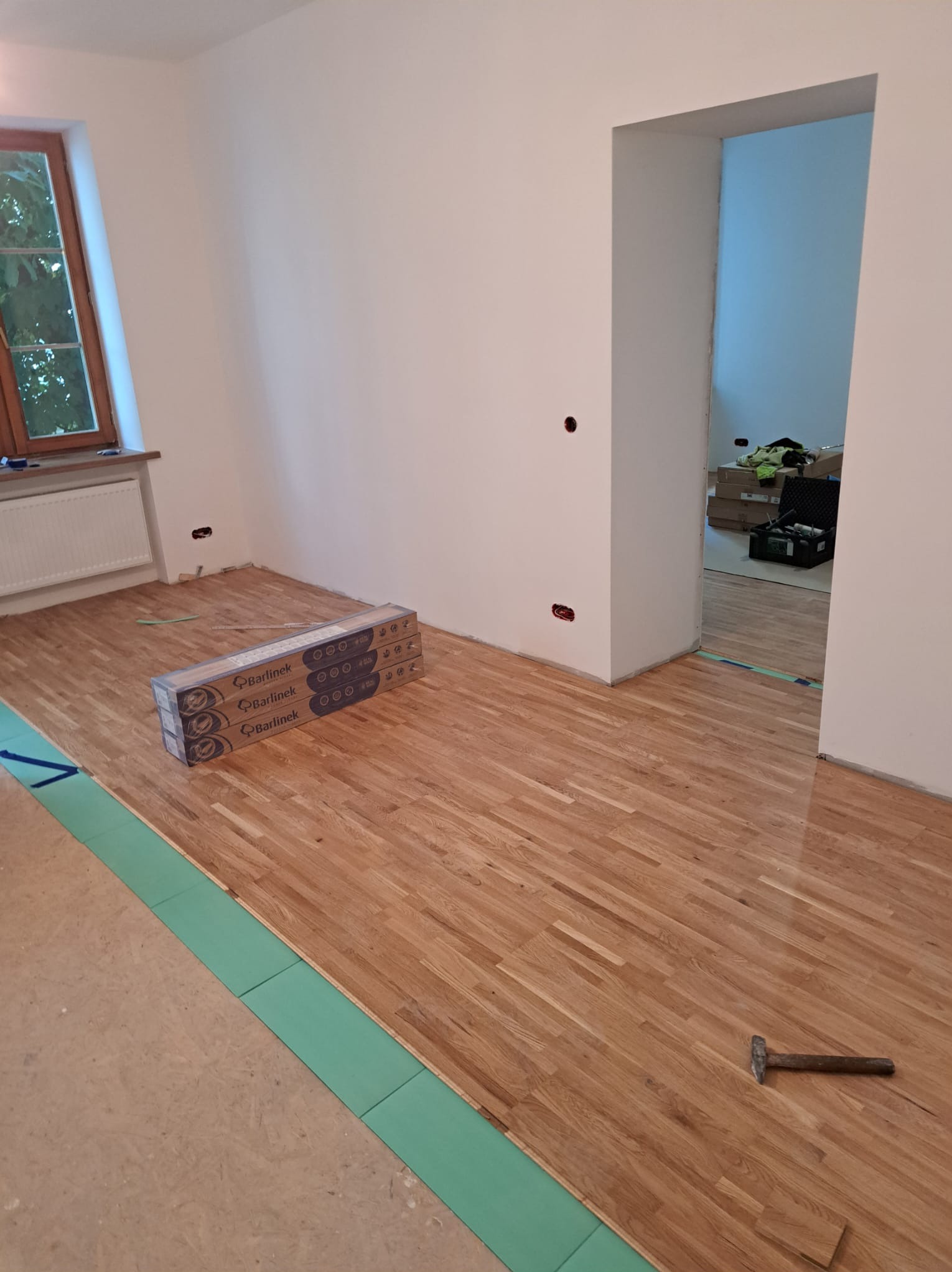 Układanie parkietu Barlinek w pokoju. Widoczny fragment podkładu, paczki paneli, młotek. Częściowo ułożona podłoga w jasnym odcieniu drewna.