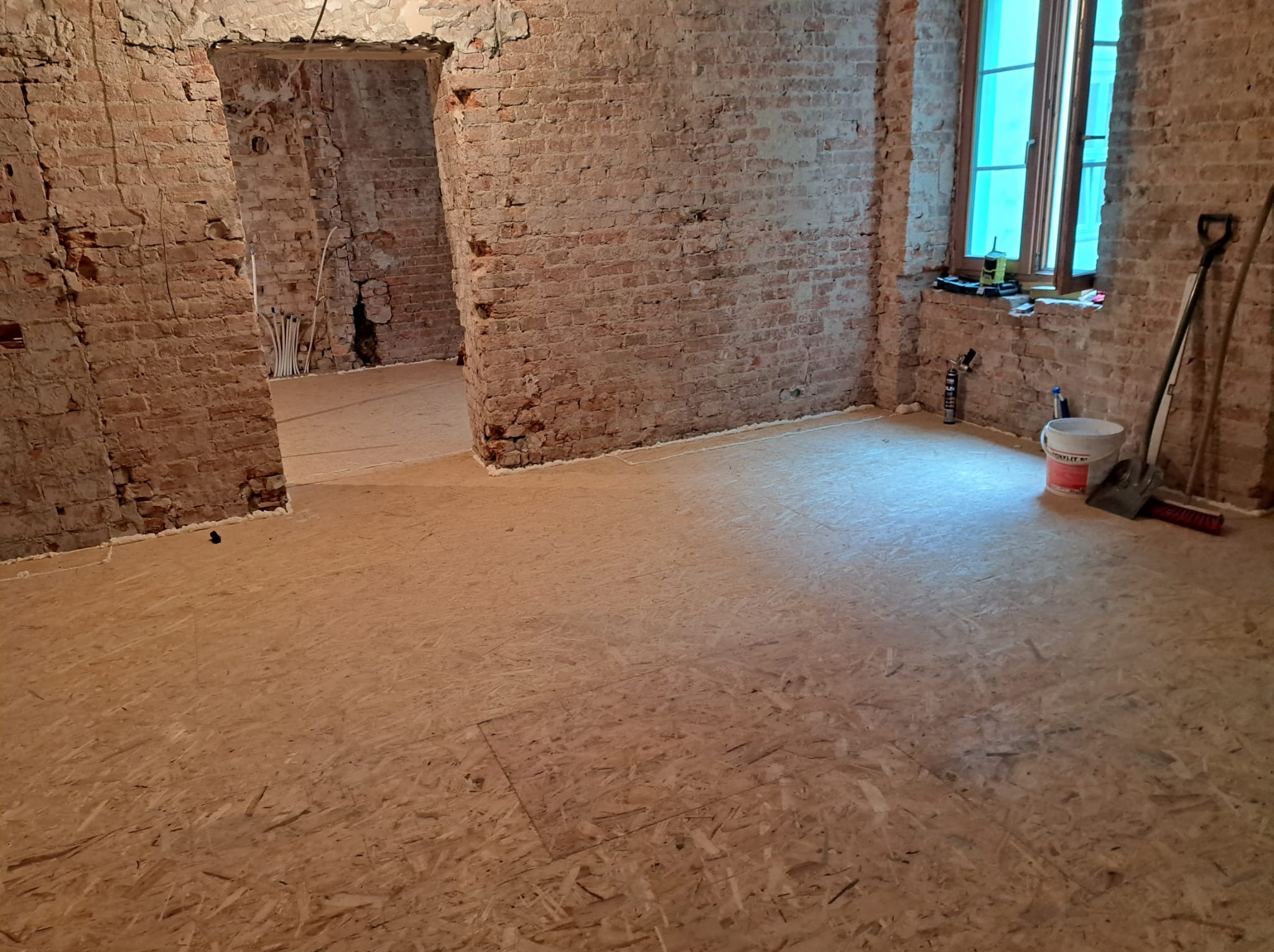 Wnętrze w trakcie remontu: surowe ceglane ściany, podłoga z płyt OSB, okno z niebieską folią. Widoczne narzędzia i materiały budowlane.