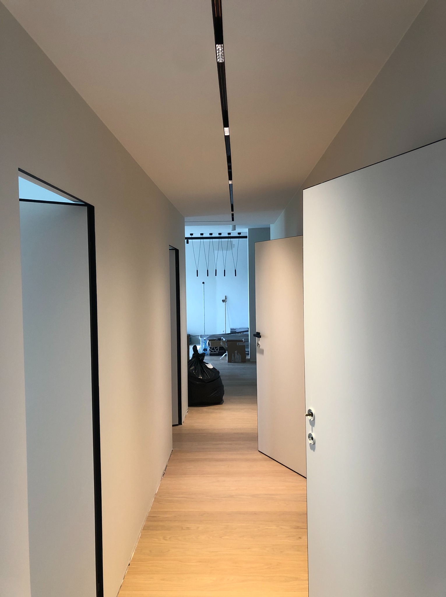 Biały korytarz po remoncie z drewnianą podłogą, czarnymi obramowaniami drzwi i nowoczesnym oświetleniem sufitowym. Widok na wnętrze z workiem i kartonami.