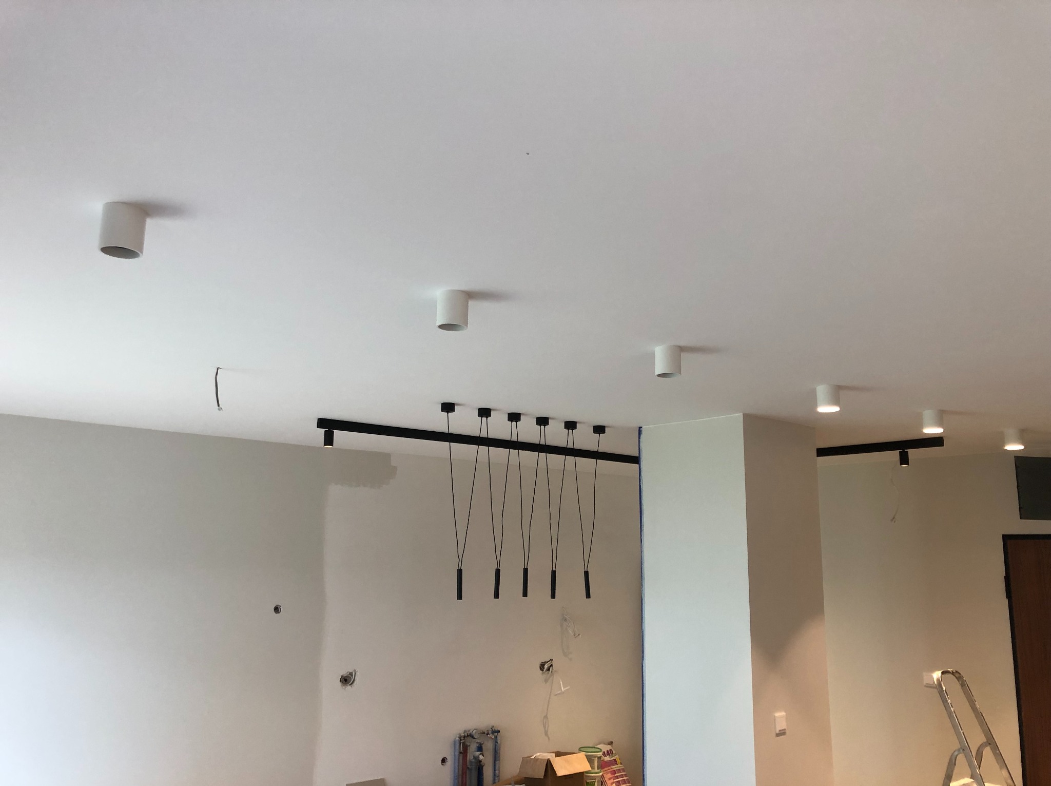 Białe wnętrze w trakcie wykończenia: zamontowane oświetlenie sufitowe, ściany przygotowane do malowania, widoczne puszki elektryczne i drabina. Minimalistyczny styl.