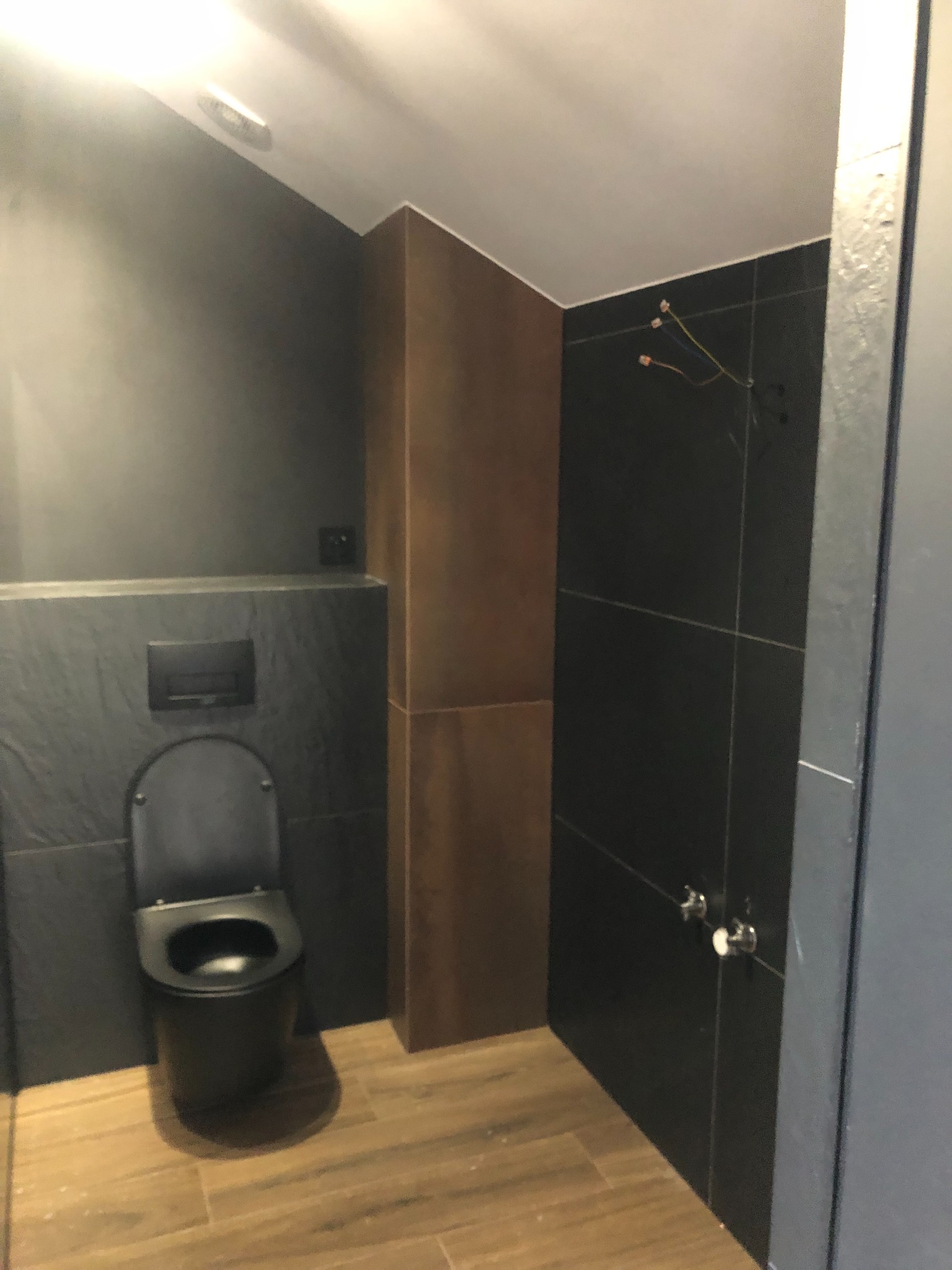 Nowoczesna łazienka z czarną armaturą i drewnopodobnymi płytkami podłogowymi. Widoczne minimalistyczne detale i oświetlenie.