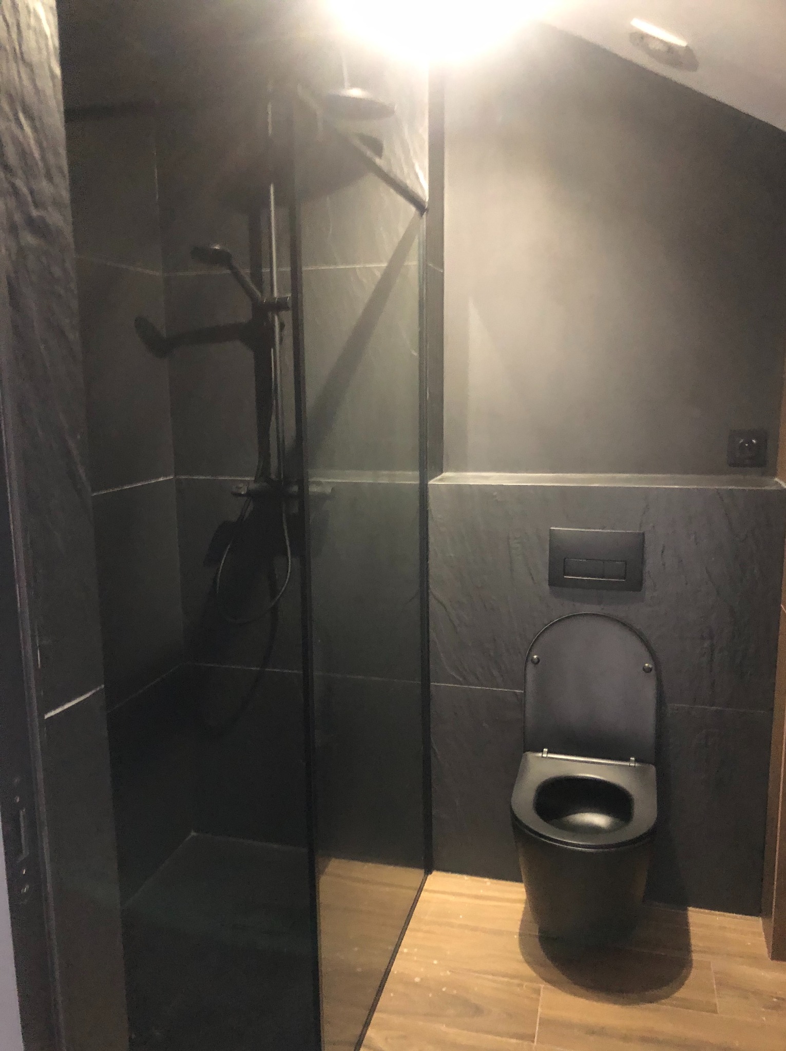 Nowoczesna, czarna łazienka z kabiną prysznicową i podwieszaną toaletą. Ściany wykończone płytkami imitującymi kamień, drewnopodobna podłoga.