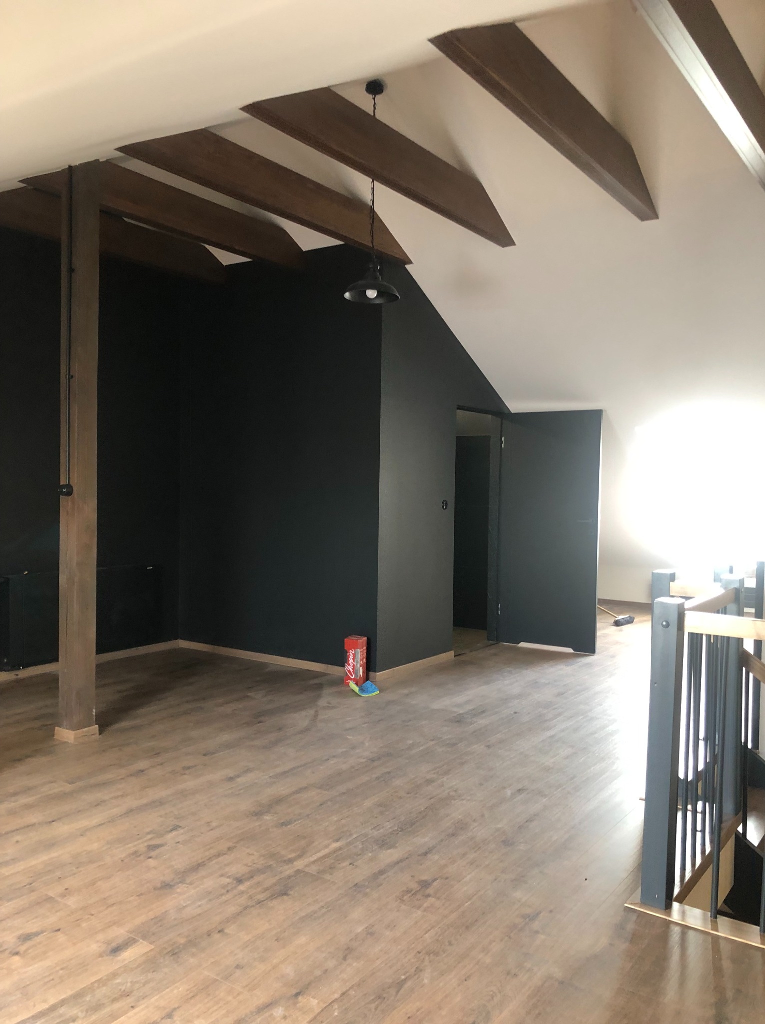 Pokój na poddaszu po malowaniu ścian na ciemny kolor, z drewnianymi belkami stropowymi, panelami podłogowymi i otwartymi drzwiami. Widoczny grzejnik i puszka farby.