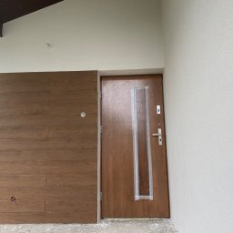 W&P TERMO-TECH - Elewacja domu z drewnianą okładziną i brązowymi drzwiami wejściowymi zabezpieczonymi folią. Ściana pomalowana na biało z wyraźną fakturą.