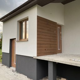 W&P TERMO-TECH - Nowoczesny dom z elewacją w odcieniach bieli i szarości, z drewnianymi akcentami przy oknach i drzwiach. Widoczne elementy wykończeniowe i fragment ogrodu.