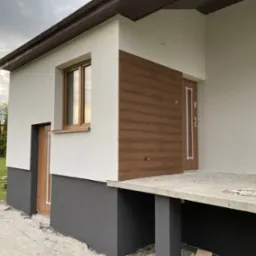 Nowoczesny dom z elewacją w odcieniach bieli i szarości, z drewnianymi akcentami przy oknach i drzwiach. Widoczne elementy wykończeniowe i fragment ogrodu.