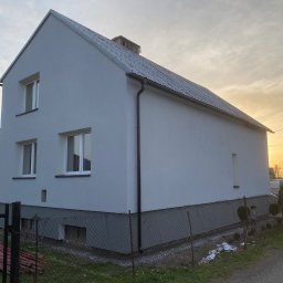 W&P TERMO-TECH - Dwukondygnacyjny dom z szarą, gładką elewacją w promieniach zachodzącego słońca. Widoczne okna, dach i fragment ogrodzenia. Minimalistyczny styl.