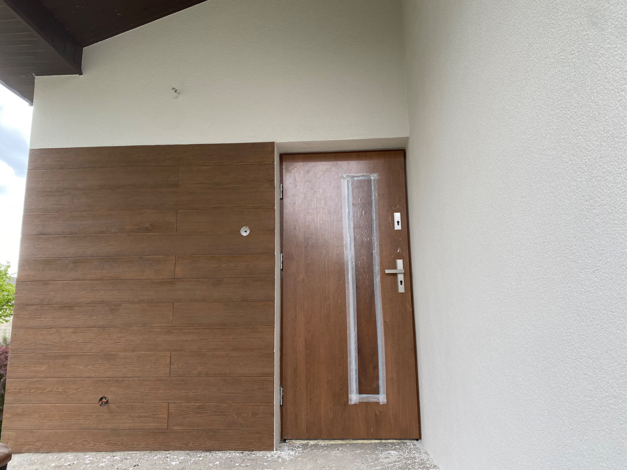 Elewacja domu z drewnianą okładziną i brązowymi drzwiami wejściowymi zabezpieczonymi folią. Ściana pomalowana na biało z wyraźną fakturą.