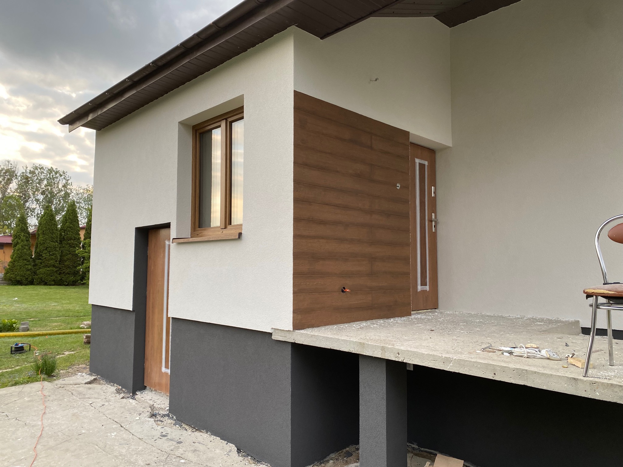 Nowoczesny dom z elewacją w odcieniach bieli i szarości, z drewnianymi akcentami przy oknach i drzwiach. Widoczne elementy wykończeniowe i fragment ogrodu.