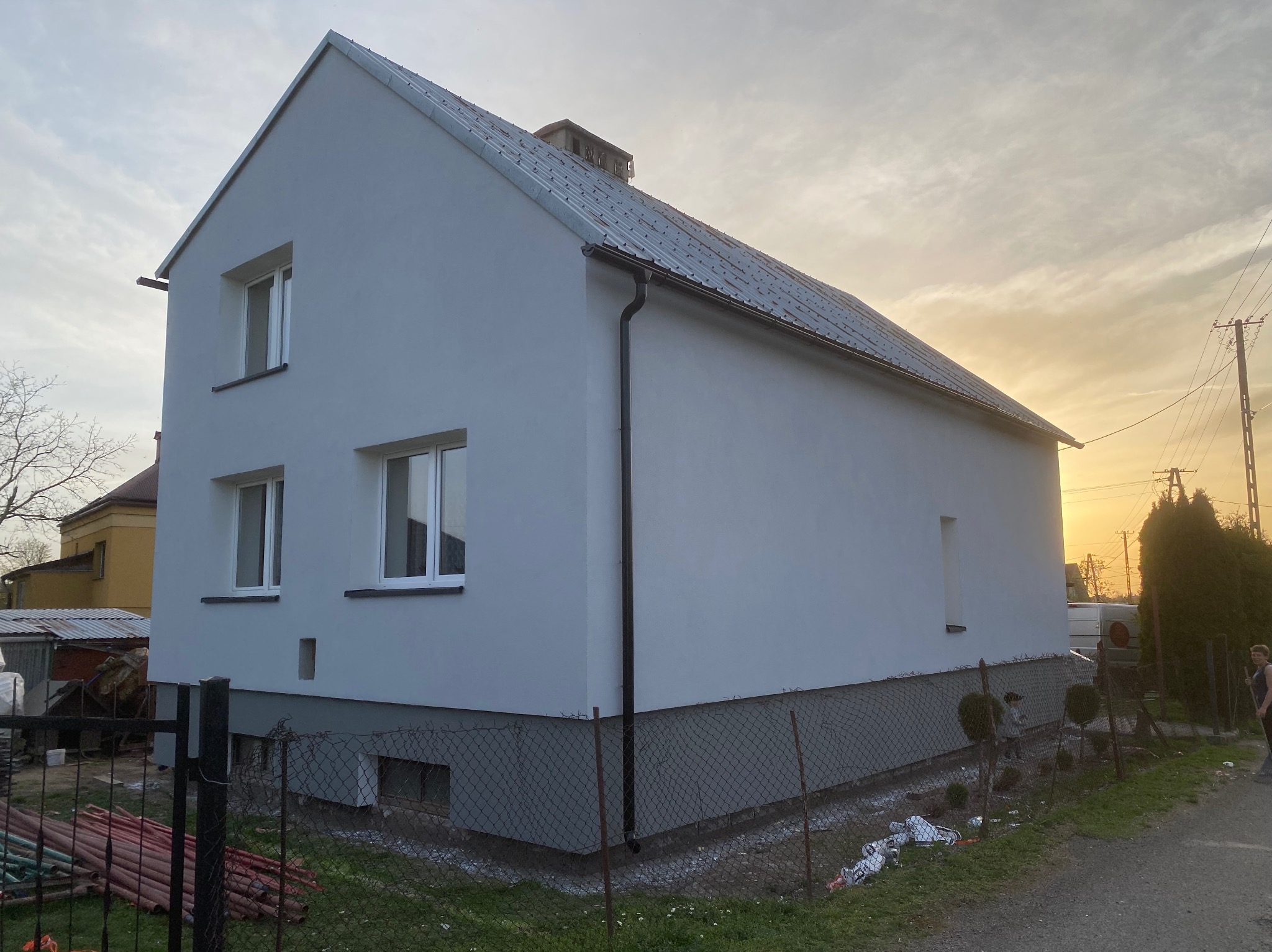 Dwukondygnacyjny dom z szarą, gładką elewacją w promieniach zachodzącego słońca. Widoczne okna, dach i fragment ogrodzenia. Minimalistyczny styl.