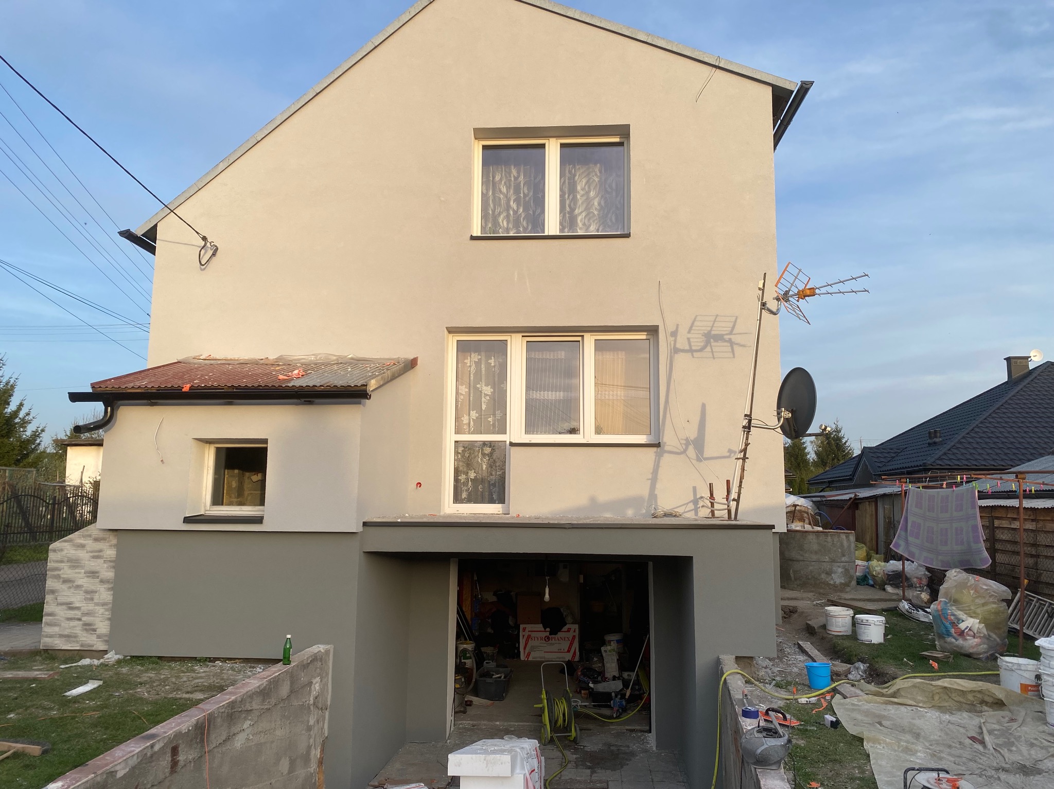 Dom po tynkowaniu z pomalowaną dolną częścią elewacji na szaro, widoczne narzędzia i materiały budowlane wokół budynku. Antena satelitarna na ścianie.
