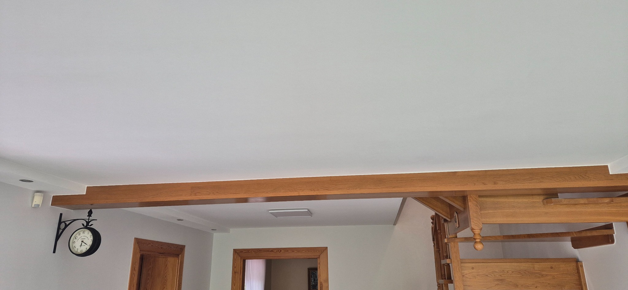Jasne wnętrze z drewnianymi elementami: belka stropowa, schody i ościeżnice drzwi. Na ścianie zegar dworcowy. Sufit biały, gładki.