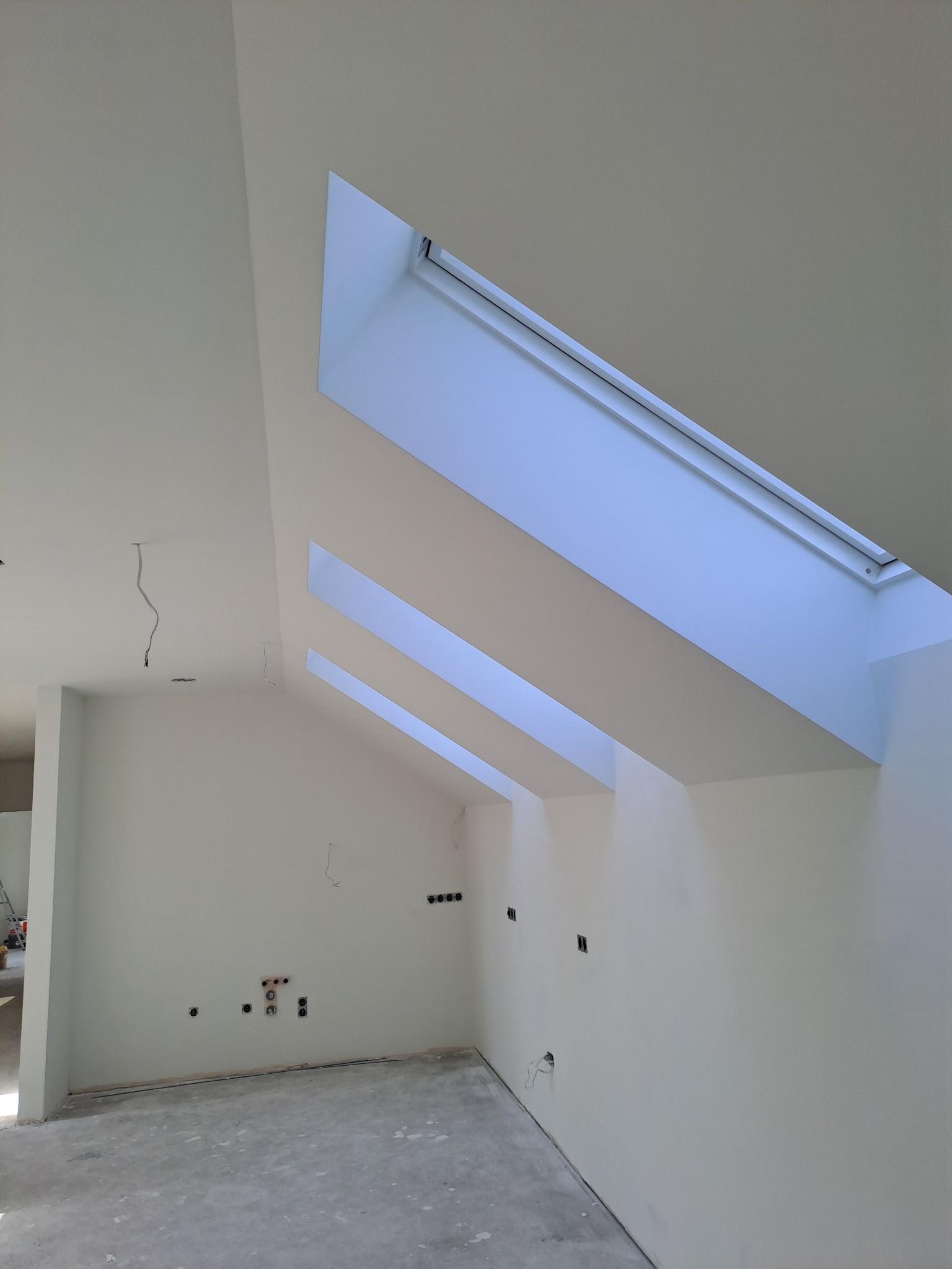 Białe wnętrze poddasza z trzema oknami dachowymi, świeżo po malowaniu. Widoczne gładkie ściany i sufit, przygotowane do dalszych prac wykończeniowych, z wystającymi przewodami.