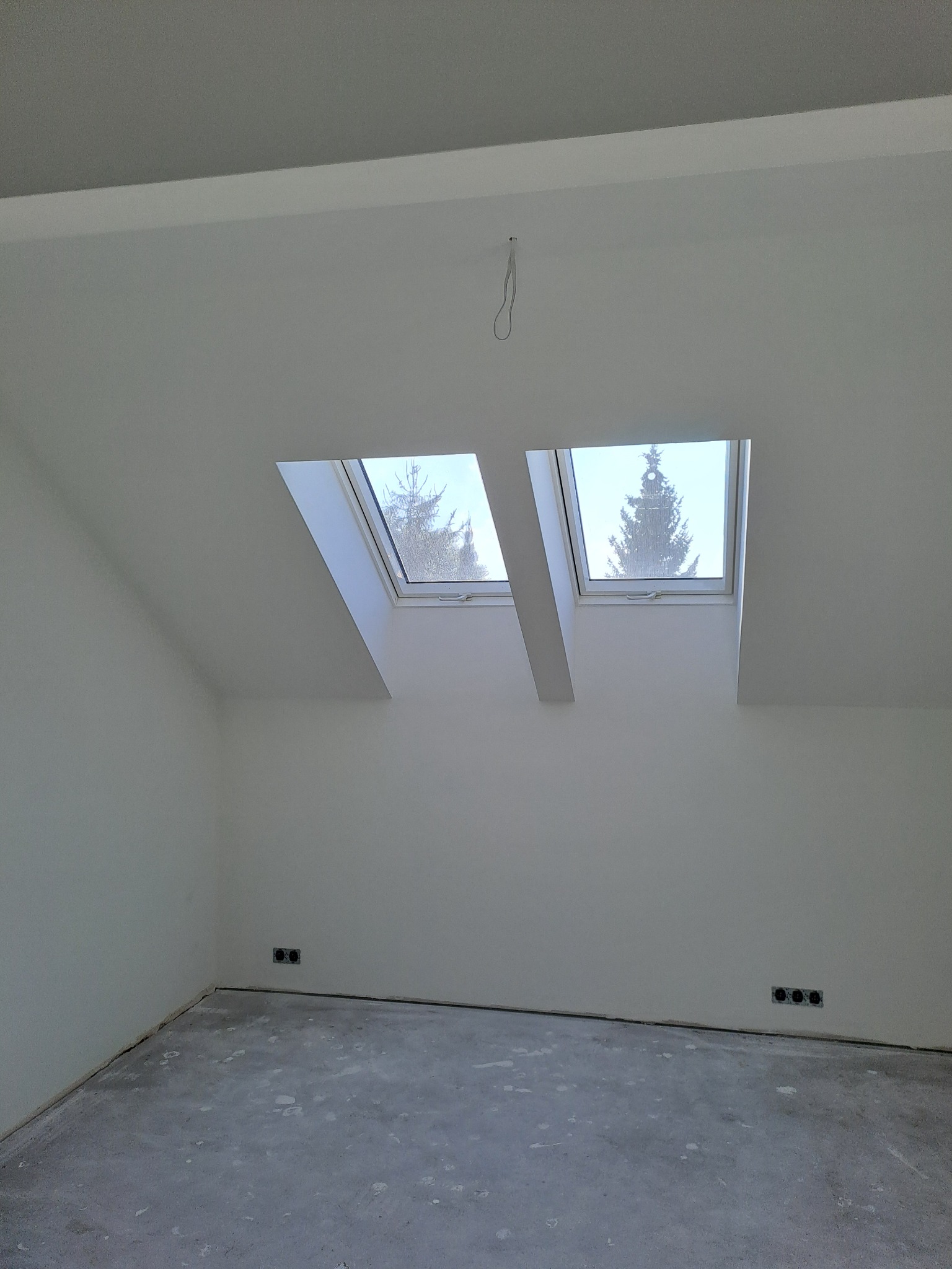 Poddasze po gładziowaniu i malowaniu na biało, z dwoma oknami dachowymi, przez które widać drzewa. Szpachlowana podłoga, widoczne gniazdka elektryczne.