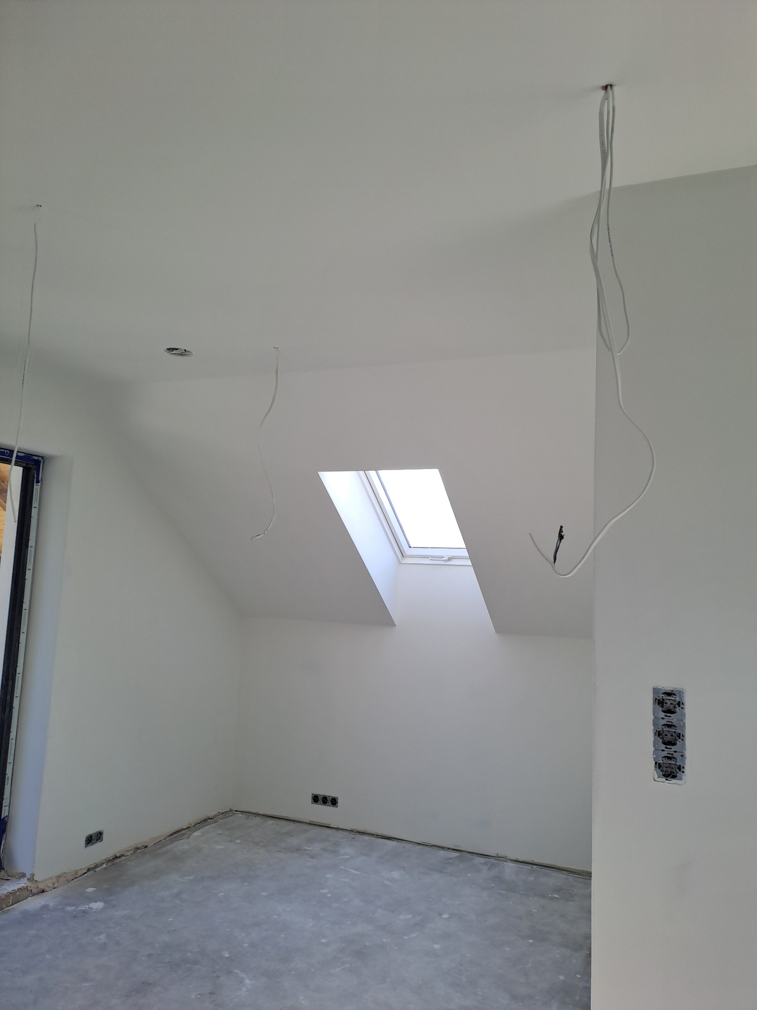 Białe, świeżo wykończone poddasze z oknem dachowym, widoczne wystające kable elektryczne i puszki gniazdek, szary beton na podłodze, ściany pomalowane na biało.