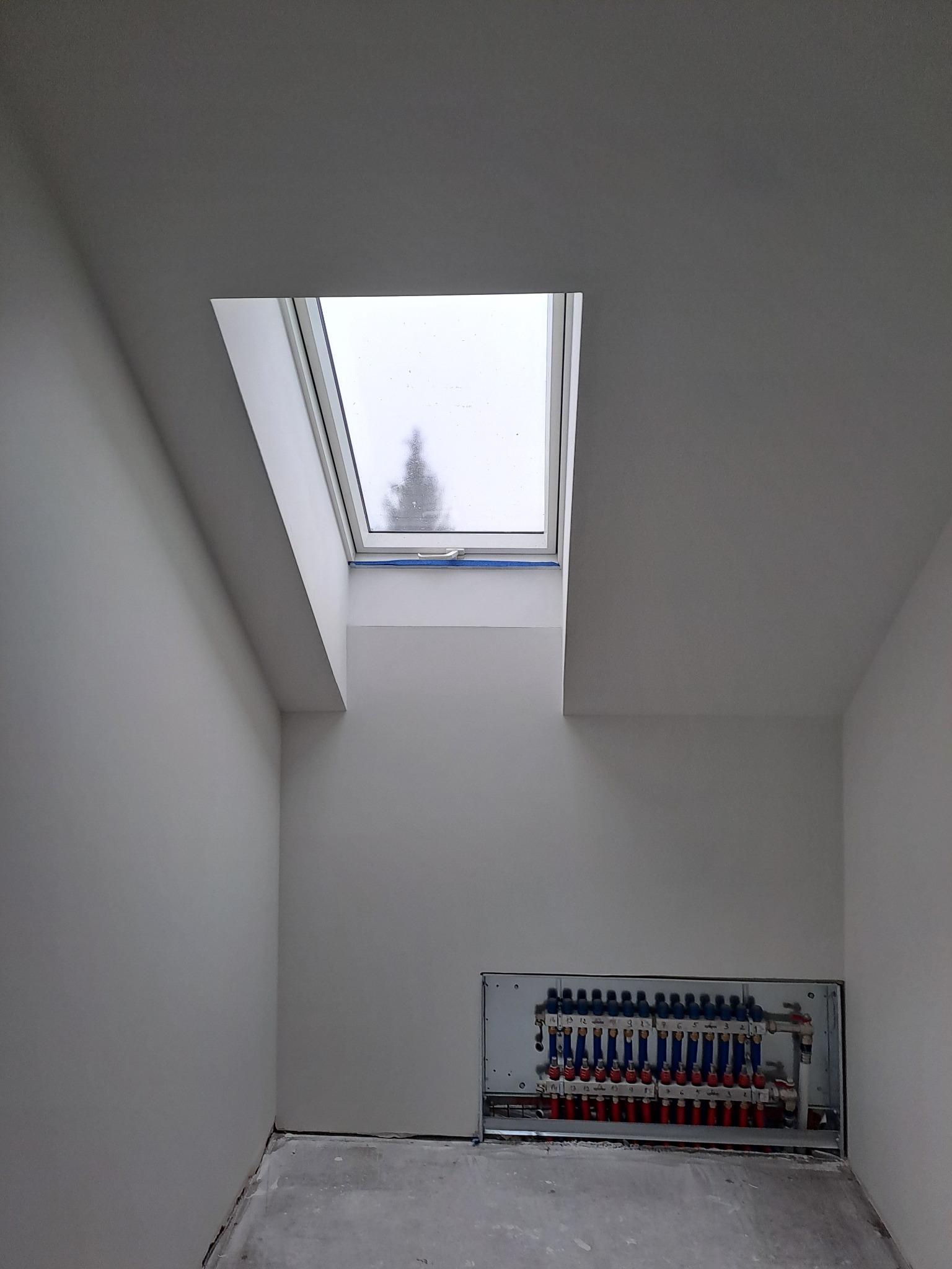 Pomieszczenie po malowaniu z oknem dachowym i rozdzielaczem ogrzewania podłogowego w ścianie. Widok na zewnątrz z drzewem. Minimalistyczny wystrój wnętrza.