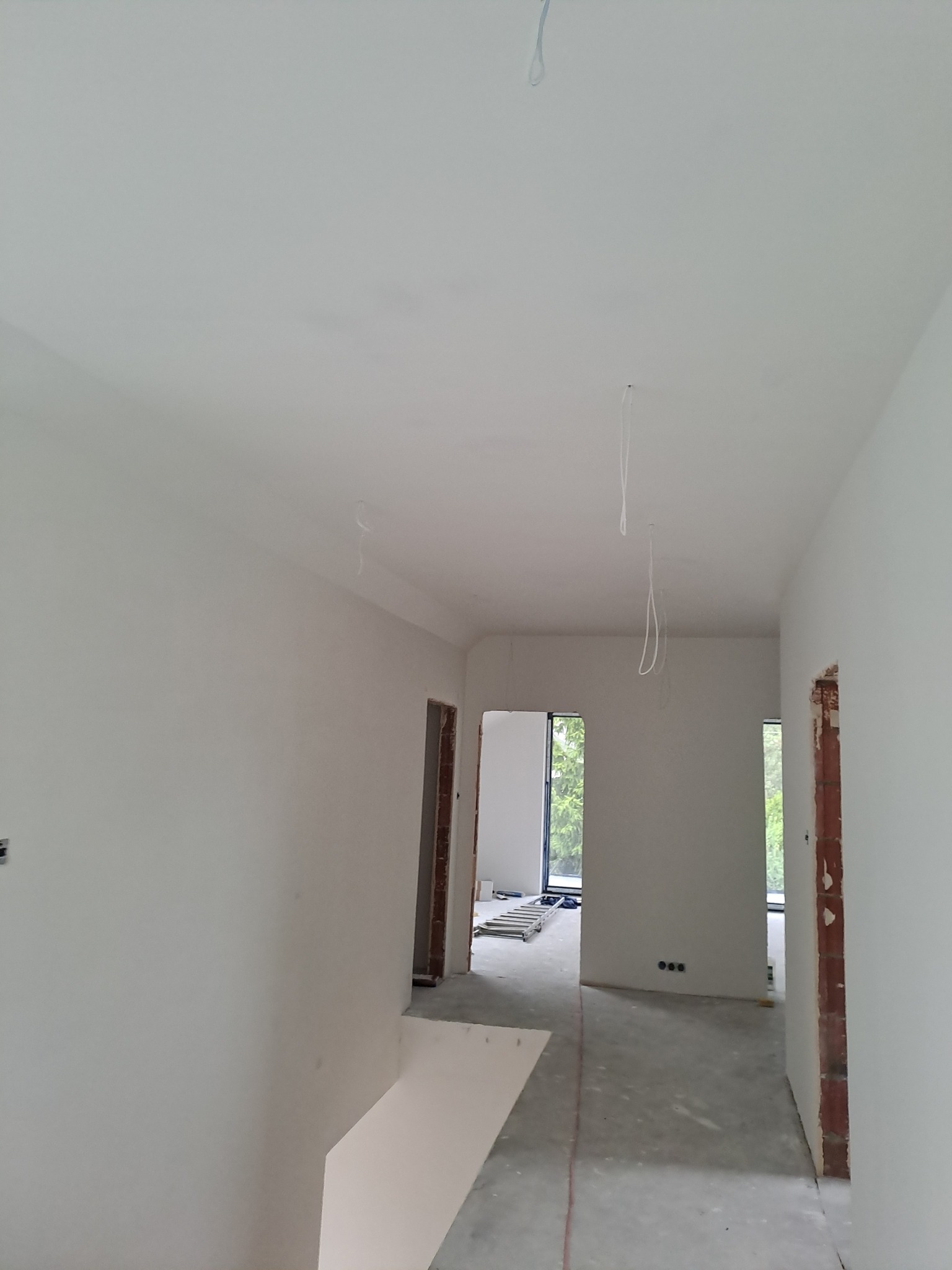 Jasne, świeżo pomalowane wnętrze w trakcie wykończenia. Widoczne gładkie ściany, sufit, surowe framugi drzwi i okien. Na podłodze widoczne ślady prac wykończeniowych.